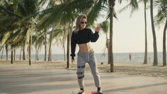 4k Video slow-motion, Follow Font camera view. An Asian woman wearing glasses plays a surf skateboard in a park with a coconut tree near the beach during the time of the sunset.