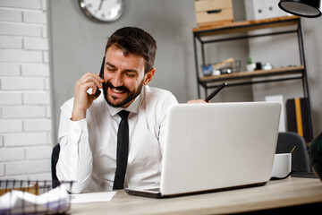 Fototapeta premium Handsome man talking to the phone at his workplace. Young businessman using laptop in his office.