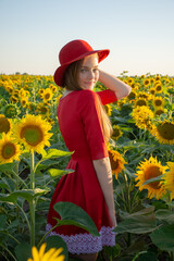 Portrait of a young girl in a red dress and hat in a field with sunflowers at sunrise