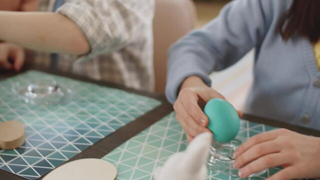 Close up shot of pretty little girl taking dyed egg from basket and putting it in glass holder while making Easter decorations together with family