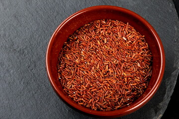 raw red rice in a clay bowl on a black stone plate. uncooked brown wild rice, top view
