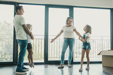 Fototapeta premium Mother, father and two daughters standing in empty living room with open balcony. Happy family relocating in new apartment or house together and watching room. Relocation and moving day concept