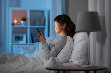 technology, internet, communication and people concept - happy smiling young asian woman with smartphone lying in bed at home at night