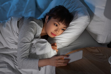 technology, internet, communication and people concept - young asian woman with smartphone lying in bed at home at night