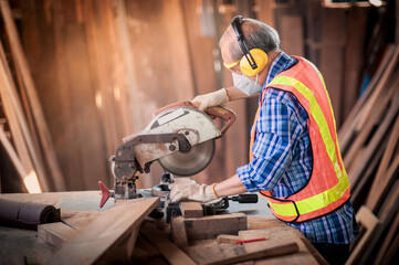 Asian elderly carpenter craftsman in carpentry shop use circular saws to cut wood board to make furniture. And wear safety equipment at work such as dust masks, gloves, noise-canceling, headphones.