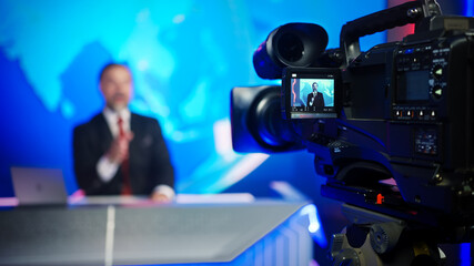 Professional TV Camera Standing in Live News Studio with Anchor seen in Small Display. Unfocused TV Broadcasting Channel with Presenter, Newscaster Talking. Mock-up Television Channel Newsroom Set - obrazy, fototapety, plakaty