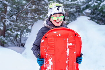 Happy boy holds a red pan sledge in his hands to ride the slide. Winter activities. Downhill. Sledging. Winter entertainment and sports. Activities for children and teenagers. Speed sled-ice © alonaphoto