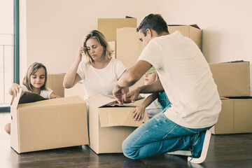 Focused parents and kids unpacking things in new apartment, sitting on floor and opening boxes. House buying or moving concept