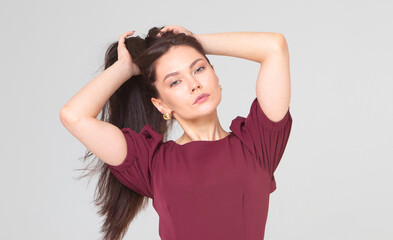 Fototapeta premium beautiful girl 25 years old, European appearance, with dark long hair, in a red dress, straightens her hair, stands on a light gray background, in the studio. Portrait of a girl, brunette. beauty