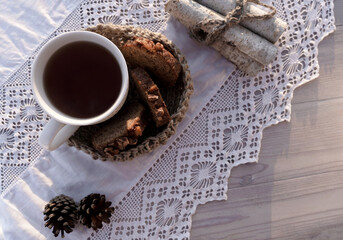Vintage rustic card with white porcelain cup of tea and cake on white wooden table decorated with cones and lacy white napkin. Winter seasonal holiday background. Food and drink concept. Top view.