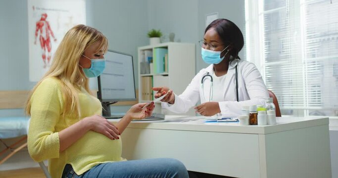 Medium shot of pregnant woman with medical mask visit african american gynecologist at hospital. Doctor in glasses giving ultrasound picture. Pregnancy woman and maternity healthcare concept.