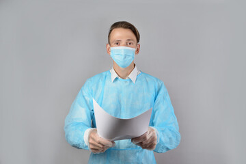 A fair-haired male surgeon in a blue uniform, wearing a medical mask and protective gloves, holds out white sheets of paper. Health care, stay at home. A young, good-looking medical worker.