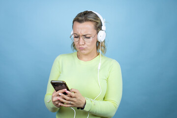 Young caucasian woman listening to music using headphones over blue background chatting with her phone