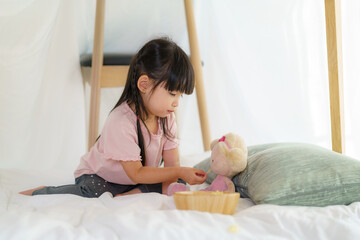 Asian cute little girl play her doll and feeding snack while sitting in a blanket fort in living...
