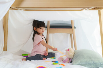 Asian cute little girl play cooking and feeding food to her doll while sitting in a blanket fort in...
