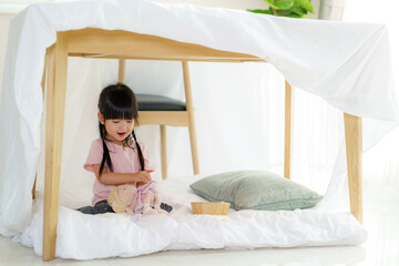 Asian cute little girl play her doll while sitting in a blanket fort in living room at home for...