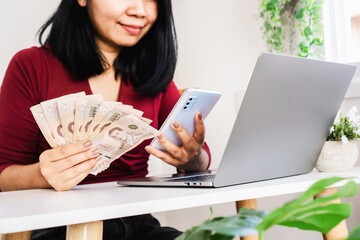 Asian woman holding Thai money using smart phone and laptop on desk, register, paying online concept