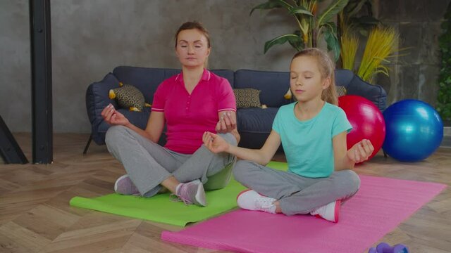 Sporty charming mother and cute preadolescent daughter meditating with eyes closed on fitness mats in yoga lotus position, peeping out each other while practicing relaxation exercise at home.