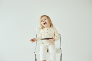 Small child girl sitting on a chair on white isolated background with smartphone in hands smiling and watching some cartoons as addiction - 410931535