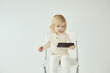 Small child girl sitting on a chair on white isolated background with smartphone in hands eating and watching some cartoons as addiction - 410931586