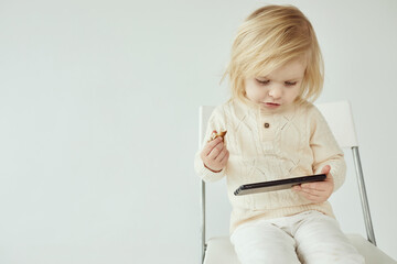 Small child girl sitting on a chair on white isolated background with smartphone in hands eating and watching some cartoons as addiction - 410931587