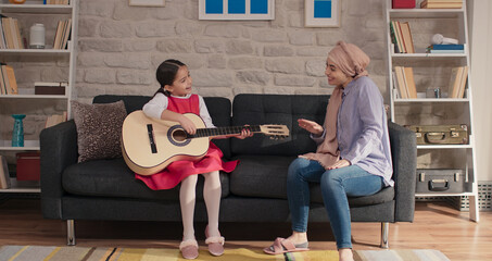 Mother wearing a turban is singing, her little daughter is having fun playing the guitar. Very happy smiling mother enjoy family leisure time at home with her little daughter. 