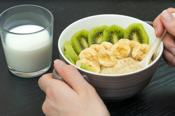 Women's hands take a spoon from a bowl of oatmeal porridge with bananas, kiwi close-up, next to a glass of milk. Concept of a healthy breakfast