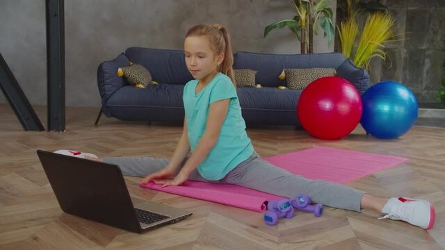 Flexible graceful lovely elementary age girl in sports clothes practicing stretching exercise, doing straddle split and communicating with fitness instructor while working out online in home interior.