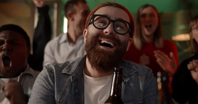 Close up of bearded man in glasses and hat drinking beer while watching sports with friends. Group of multiethnic young people celebrating and rejoicing while spending time in pub