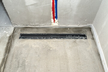 A modern black linear shower drain installed in the shower floor, visible water pipes in the wall.