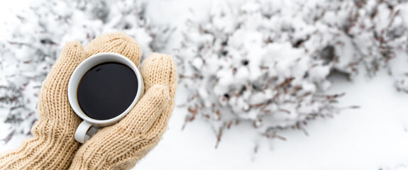 Female hands in woolen knitted gloves holding cup of warm black coffee outdoors on snowy background. Top view of hands and coffee mug. Happy, frosty winter morning