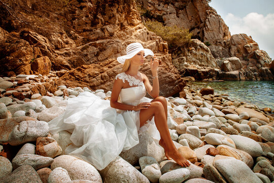 Beautiful sexy blonde woman in wedding dress with white hat and long hair and tanned body sitting at the seashore.