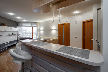 Modern interior of kitchen in luxury flat. New kitchen set with light wooden counter. Bar stool. Living zone with couch. Wooden shelf with drawers.