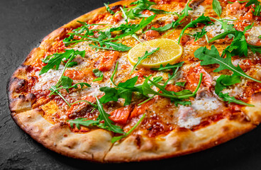 Pizza with Mozzarella cheese, salmon fish, tomato sauce, arugula, lemon. Italian pizza on Dark grey black slate background