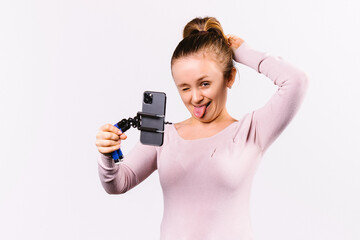 The blonde girl winks and shows her tongue while making a selfie on her smartphone. White background and playful mood.