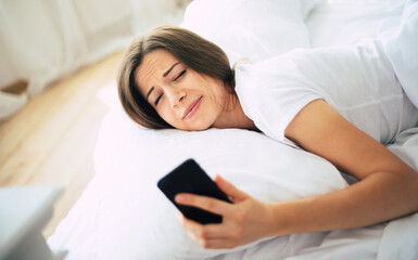Tired unhappy serious young woman during awakening in the morning in bed with smartphone in hand