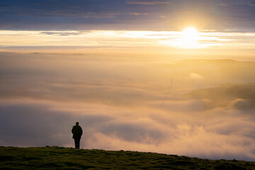 Silhouette of one lone man stood on edge of mountain peak top above clouds as fog mist and cloud inversion gathers watching the sun rise with a beautiful orange sunlight sky 