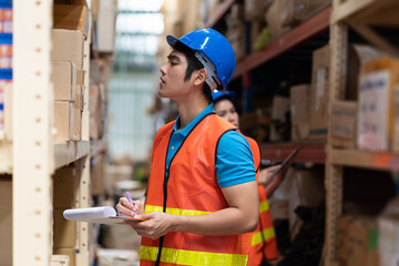 Asian male and female warehouse worker working with clipboard for checking products on shelves shelf pallet in industry storage warehouse. Wearing safety vest and helmet