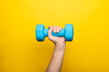 Woman's hand holding blue dumbbell isolated on yellow background. Home workout, fitness, and activity. Sport and healthy lifestyle concept with copy space.