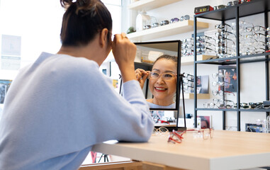 Selective focus at the mirror.  Senior elder Asian women smile and look at the mirror while choosing beautiful glasses frame trial inside of optical shop, store.  Eye sight for elder, healthcare.