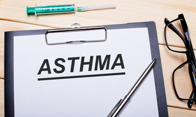 The word ASTHMA is written on a white piece of paper next to black-rimmed glasses, a pen and a syringe. Medical concept