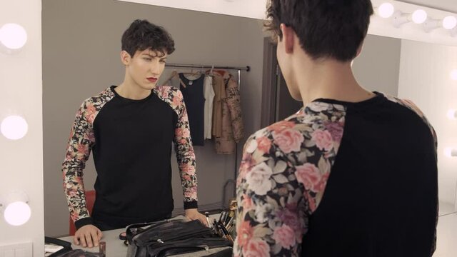 Transgender man with makeup looks in the mirror doubtfully. Unsure androgynous guy. Gender identity concept 4k