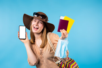 Caucasian hipster young girl wearing summer hat, handbag and sunglasses showing mockup white phone screen to camera. Blonde holds passport and international certificate of vaccination. Safe tour
