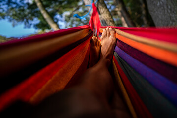 Close-up with the feet of a man enjoying a holiday in nature lying in a colorful hammock in a forest in a green city park.