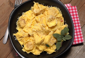 Cappelletti di pasta all'uovo ripieni di formaggio e speck tirolese aromatizzati con burro e salvia in un piatto scuro su fondale di legno.