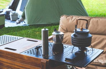 Vintage outdoor black kettle and coffee grinder on camping table with brown armchair on green lawn...