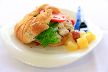 A close-up image of a croissant sandwich with chicken, lettuce, tomato, pineapple, and grapes served on a white plate. Focus is purposely narrow and at the front face of the image. 
