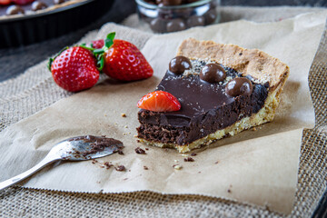 Eaten chocolate tart or pie slice topped with strawberries, chocolate candy and almonds. Delicious gluten-free homemade crust or tartlet on a dark table and brown cloth. Dessert spoon in background.