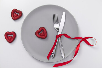 Fototapeta na wymiar Valentine's day festive table setting, flat lay with red heart shape on gray plate, fork, knife and red ribbons on white table. Love dating concept. Place your text and copy space. High quality photo