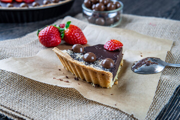 Eaten chocolate tart or pie slice topped with strawberries, chocolate candy and almonds. Delicious gluten-free homemade crust or tartlet on a dark table and brown cloth. Dessert spoon in background.